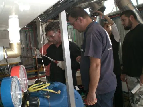 Lucrare practică la o instalație de conducte: un participant cu ochelari de protecție manevrează o trusă de lipit, urmărit de ceilalți cursanți