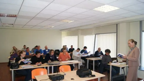 Participanți așezați la mese, răsfoind broșurile de curs, în timp ce lectoarea le distribuie din partea dreaptă a sălii