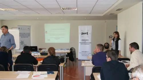 Prezentare într-o sală de curs: lector în picioare lângă ecranul de proiecție și un roll-up AGFR, cu participanți așezați la mese