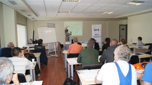 Sală de curs cu flipchart și o schemă proiectată pe perete, cu participanți așezați la mese