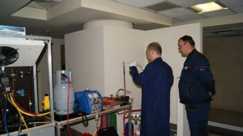 Doi tehnicieni în salopetă albastră la un stand didactic cu agregat frigorific, butelie și baterie de manometre; unul dintre ei citește o valoare
