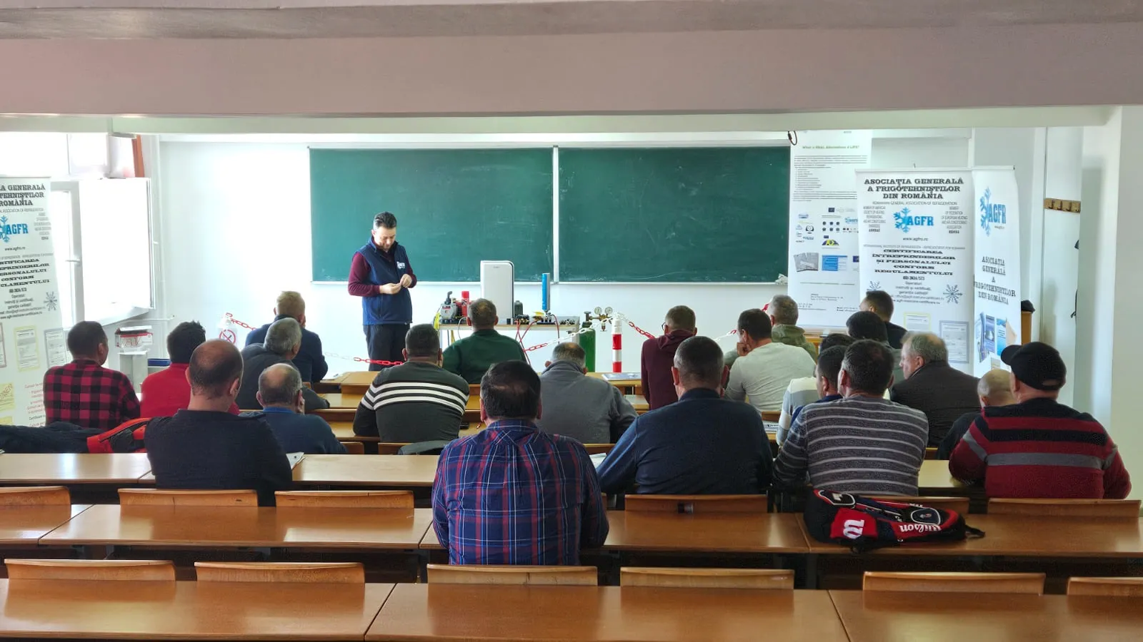 Amfiteatru cu participanți așezați în bănci, un instructor lângă un stand cu echipamente frigorifice și roll-up-uri AGFR despre certificarea conform Regulamentului (UE) 2024/573